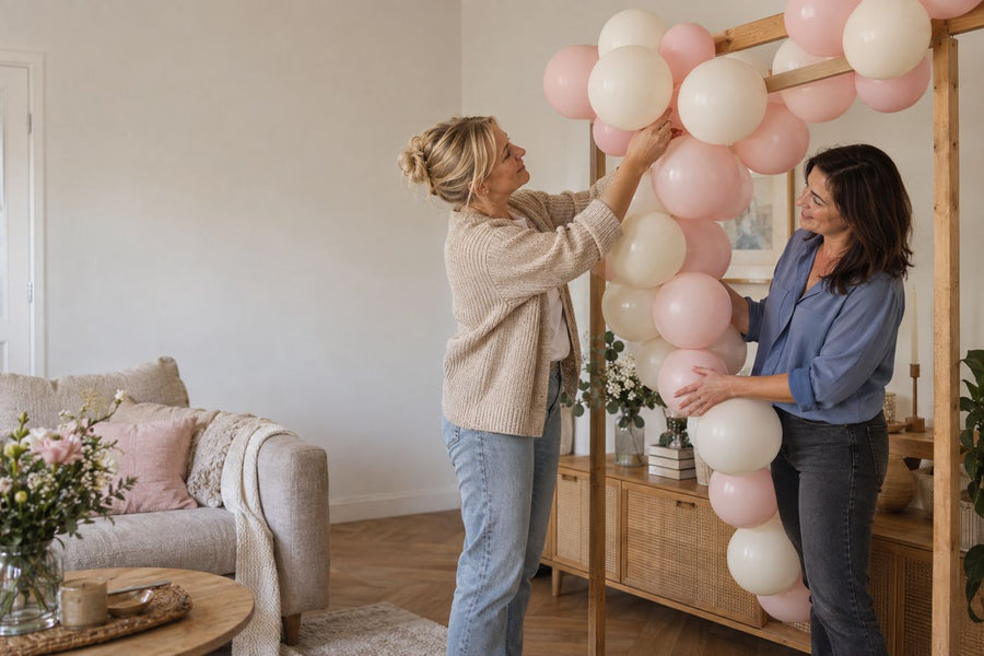 Twee vrouwen werken aan een roze en ivoorkleurige ballonnenslinger in een woonkamer.