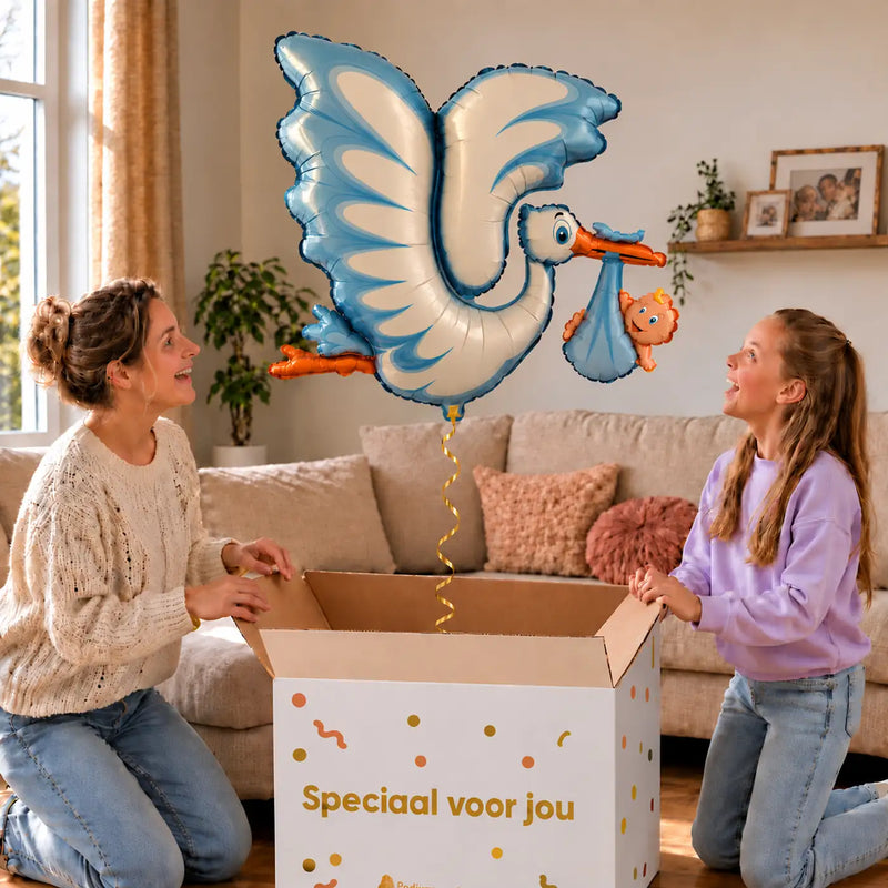 Blauwe Ooievaar Helium XL Ballon zweeft boven een geopende cadeaudoos bij een moeder en meisje in een warme woonkamer.
