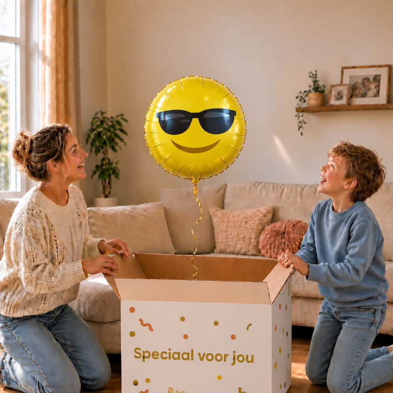 Moeder en jongen bekijken een gele emoji-ballon van 45 cm met zwarte zonnebril boven een geopende cadeaudoos.