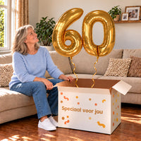 60 Jaar Cijfer Ballonnen Goud met twee heliumgevulde cijferballonnen en een jarige in een warme woonkamer