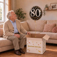 Man van 80 jaar pakt een zwarte, zilveren en gouden 80-jaarheliumballon uit een feestelijke doos.