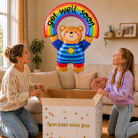 Beer Get Well Soon Regenboog XL zweeft boven een geopende cadeaudoos bij een moeder en meisje in een warme woonkamer.