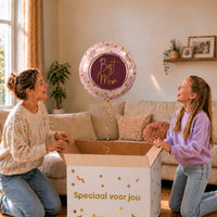 Moeder en meisje bekijken een roze Best Mom-ballon van 45 cm met gouden hartjes boven een geopende cadeaudoos.