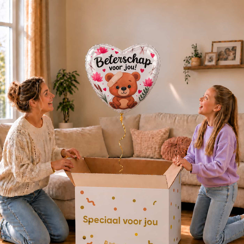 Moeder en meisje bekijken een hartvormige Beterschap voor jou-ballon van 46 cm met een beertje, pleister en bloemen boven een geopende cadeaudoos.
