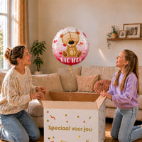 Moeder en meisje bekijken een Big Hug-ballon van 45 cm met een beertje en rode hartjes boven een geopende cadeaudoos.