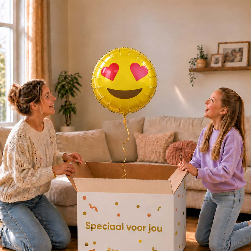 Moeder en meisje bekijken een gele emoji-ballon van 45 cm met rode hartjesogen boven een geopende cadeaudoos.