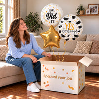 Een blije geslaagde met Geslaagd Trosje Helium Ballonnen boven een open Speciaal voor jou-doos in een warme woonkamer.