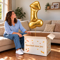 Een vrouw kijkt blij naar Gouden Piemel Helium XL Ballon boven een grote Speciaal voor jou-doos in een warme woonkamer.
