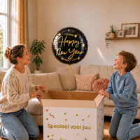 Moeder en jongen kijken naar een zwarte Happy New Year heliumballon met gouden letters en sterren boven een geopende cadeaudoos.