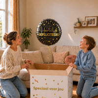 Moeder en jongen bekijken een zwarte Hartelijk Gefeliciteerd heliumballon met gouden sterren boven de geopende cadeaudoos.