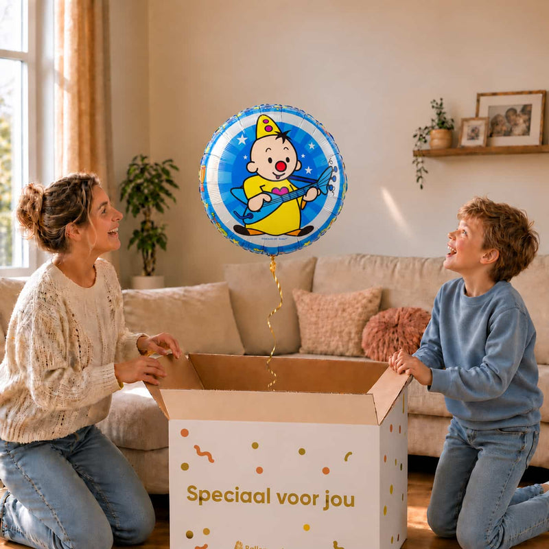 Moeder en jongen bekijken een blauwe Bumba heliumballon met Bumba en zijn gitaar boven de geopende cadeaudoos.