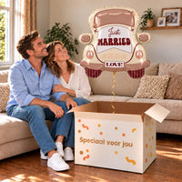Een volwassen stel kijkt blij naar Just Married Auto XL Ballon boven een grote Speciaal voor jou-doos in een warme woonkamer.