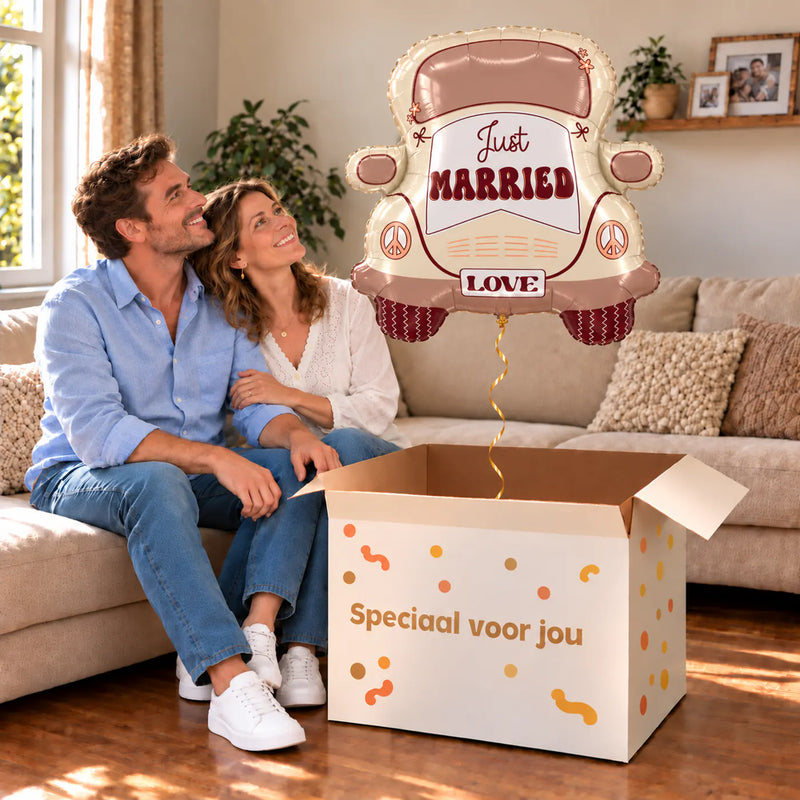 Een volwassen stel kijkt blij naar Just Married Auto XL Ballon boven een grote Speciaal voor jou-doos in een warme woonkamer.