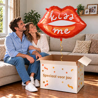 Een volwassen stel kijkt blij naar Kiss Me Helium XL Ballon boven een grote Speciaal voor jou-doos in een warme woonkamer.