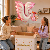 Roze Ooievaar XL Ballon zweeft boven een geopende cadeaudoos bij een moeder en meisje in een warme woonkamer.